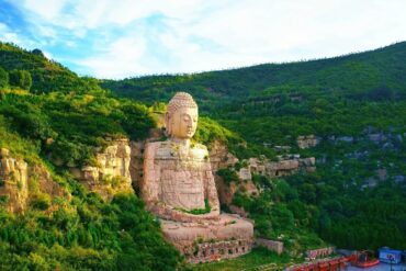 Massive stone Buddha statue carved into a rocky hillside surrounded by dense green foliage and cliffs under a blue sky, overlooking a valley.Mengshan Giant Buddha,Taiyuan, Shanxi