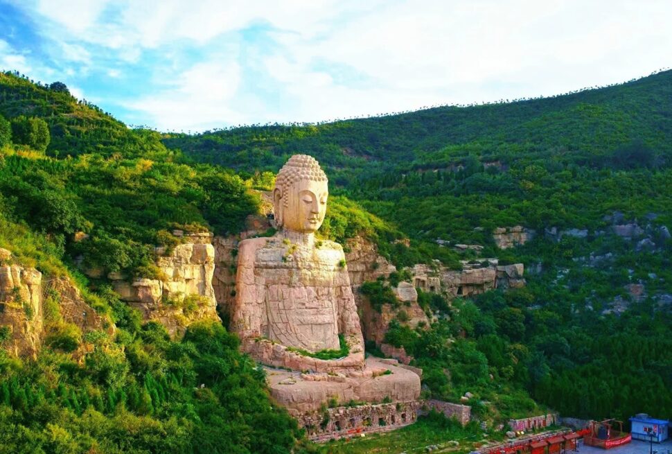 Massive stone Buddha statue carved into a rocky hillside surrounded by dense green foliage and cliffs under a blue sky, overlooking a valley.Mengshan Giant Buddha,Taiyuan, Shanxi