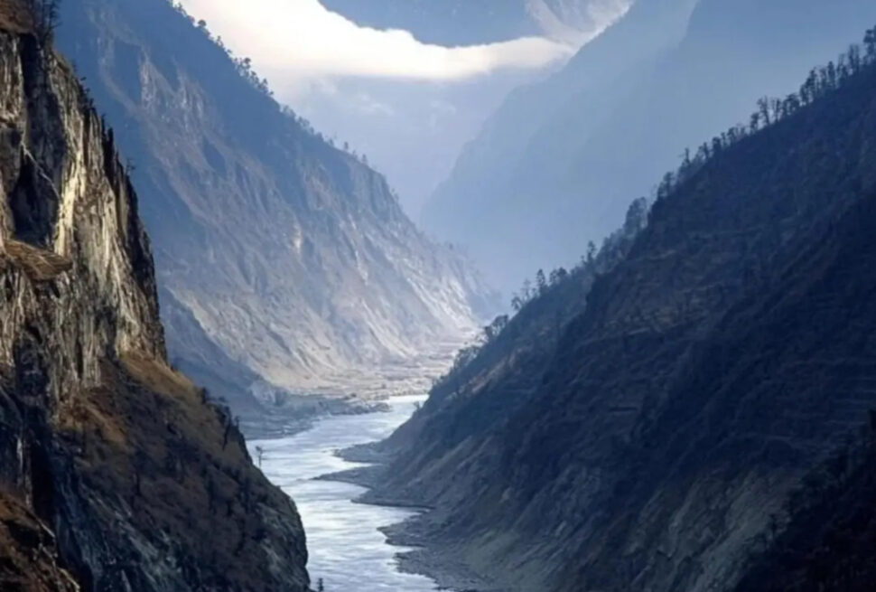 Narrow canyon with a glacial river winding between steep rocky cliffs while snow-capped mountains rise in the distance under a blue sky.