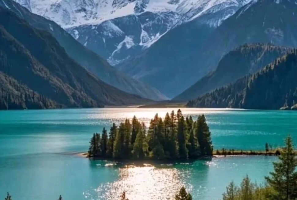 Turquoise lake with a tree-covered island, surrounded by forested mountains and snow-capped peaks in the distance
