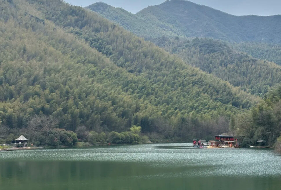 Tranquil lake surrounded by forested hills and distant blue mountains, with a docked boat and a pavilion on the left shore.