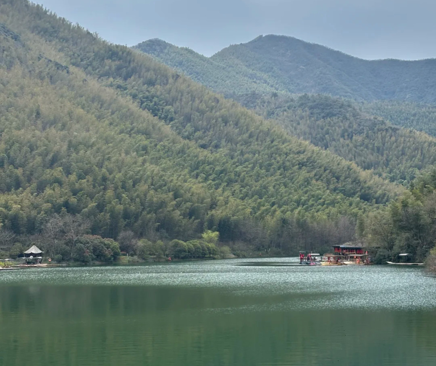 Tranquil lake surrounded by forested hills and distant blue mountains, with a docked boat and a pavilion on the left shore.