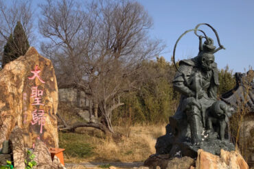 Bronze statue of a mythic Chinese warrior holding a circular weapon, perched on rocks beside a rock with red Chinese inscriptions in a park setting with leafless trees nearby.