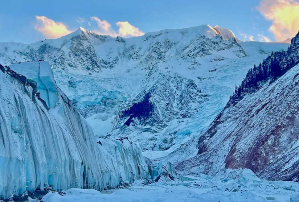 Snowy glacier with tall ice seracs and jagged mountain peaks under a blue sky at sunset.
