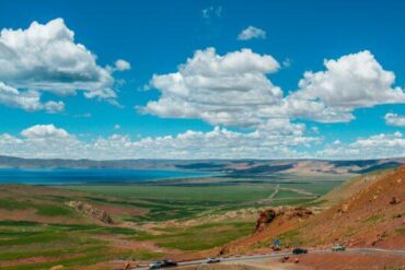Wide valley landscape: red rocky hillside on the right, winding road at the foreground, green plains, a lake in the distance under a blue sky with white clouds.