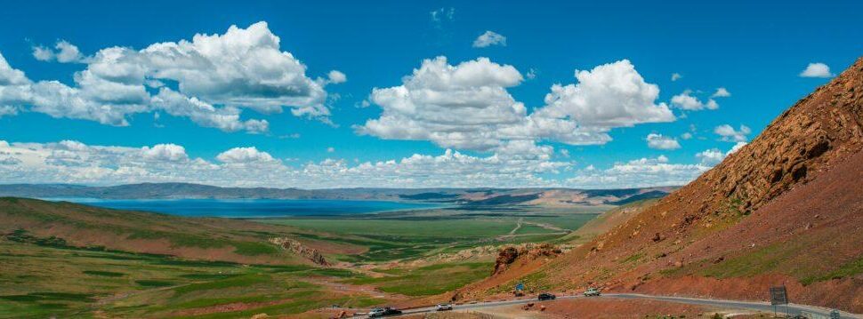 Wide valley landscape: red rocky hillside on the right, winding road at the foreground, green plains, a lake in the distance under a blue sky with white clouds.