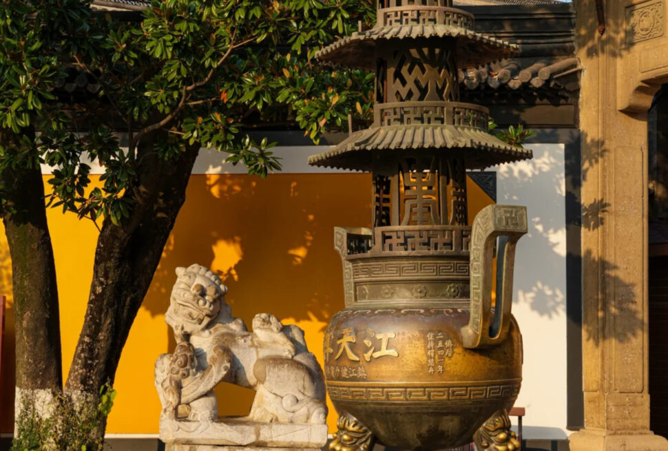 Bronze ceremonial censer with a pagoda lid on ornate tripod legs beside a stone lion statue, set against a yellow wall and leafy tree.