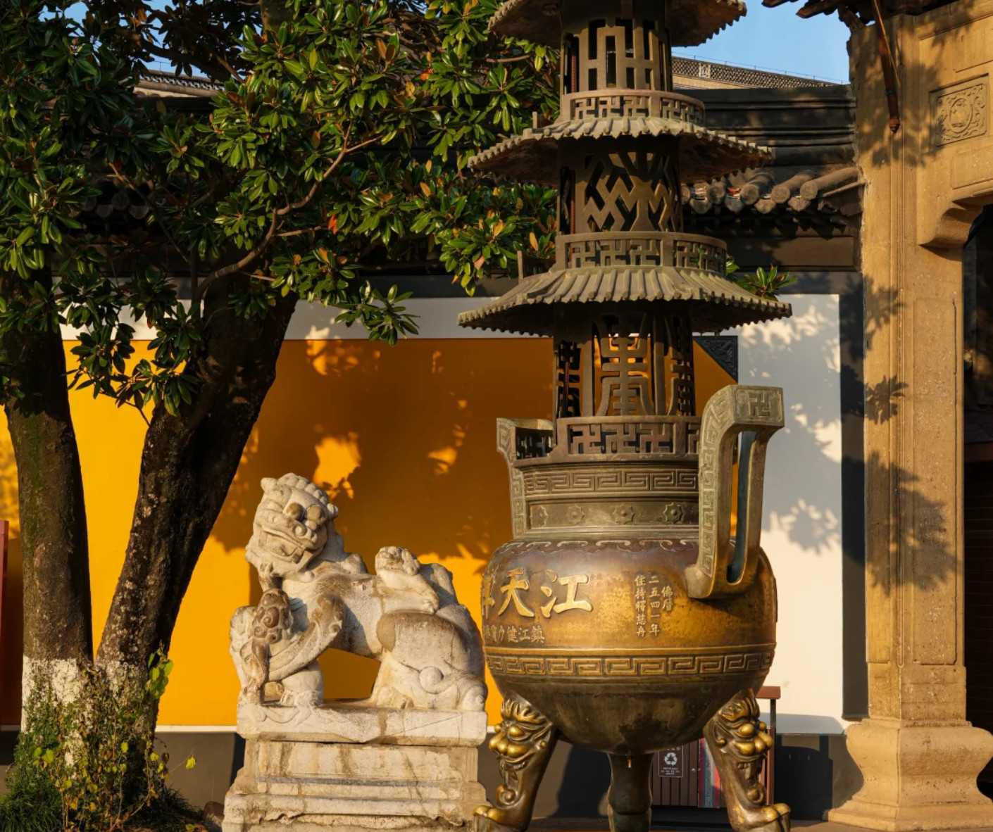 Bronze ceremonial censer with a pagoda lid on ornate tripod legs beside a stone lion statue, set against a yellow wall and leafy tree.