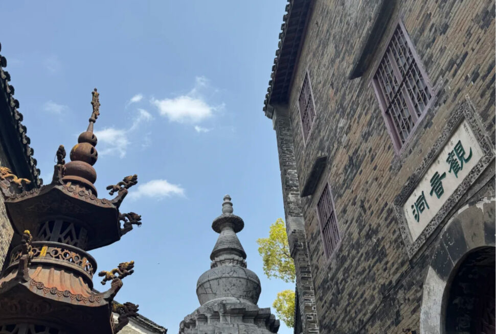Temple courtyard with a gray stone pagoda center-right, an ornate bronze structure on the left, and a brick building with a Chinese inscription on the right.