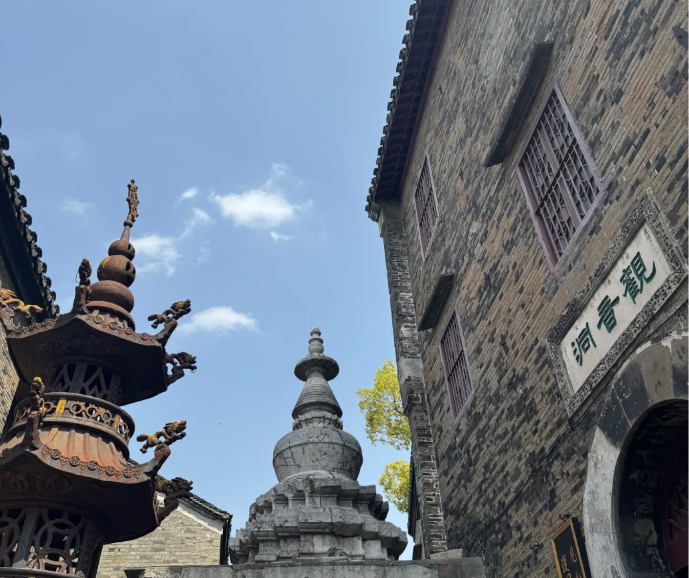 Temple courtyard with a gray stone pagoda center-right, an ornate bronze structure on the left, and a brick building with a Chinese inscription on the right.