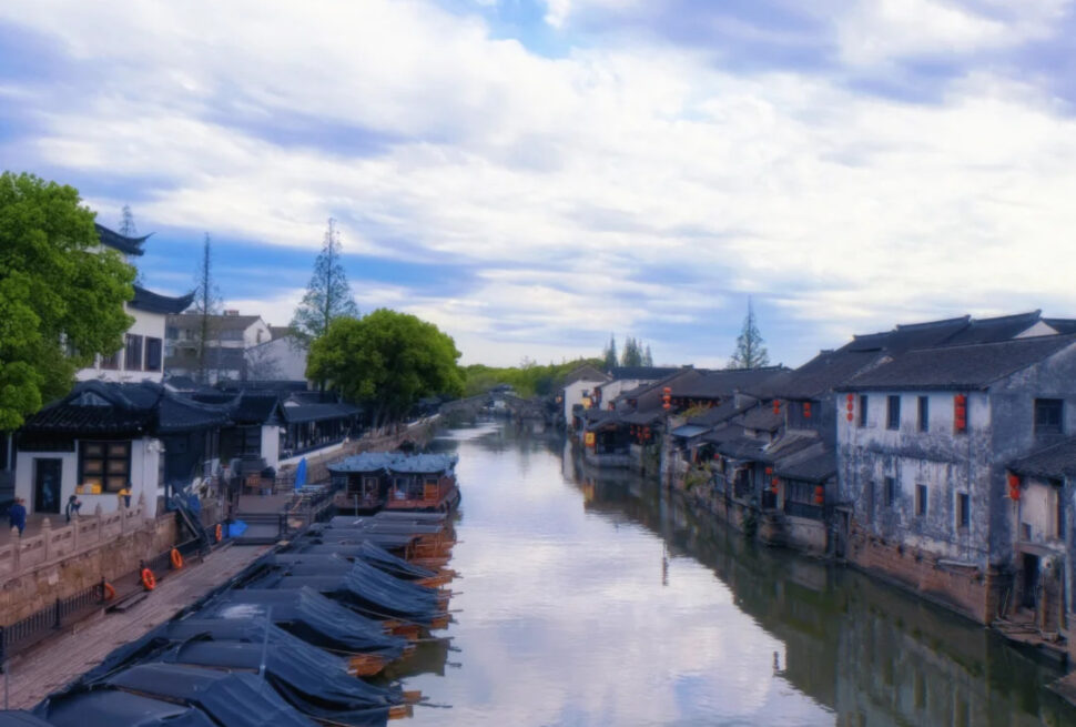 A tranquil canal lined with traditional white-walled buildings and black-tiled roofs, moored boats with blue covers, and red lanterns along the waterfront under a cloudy sky.