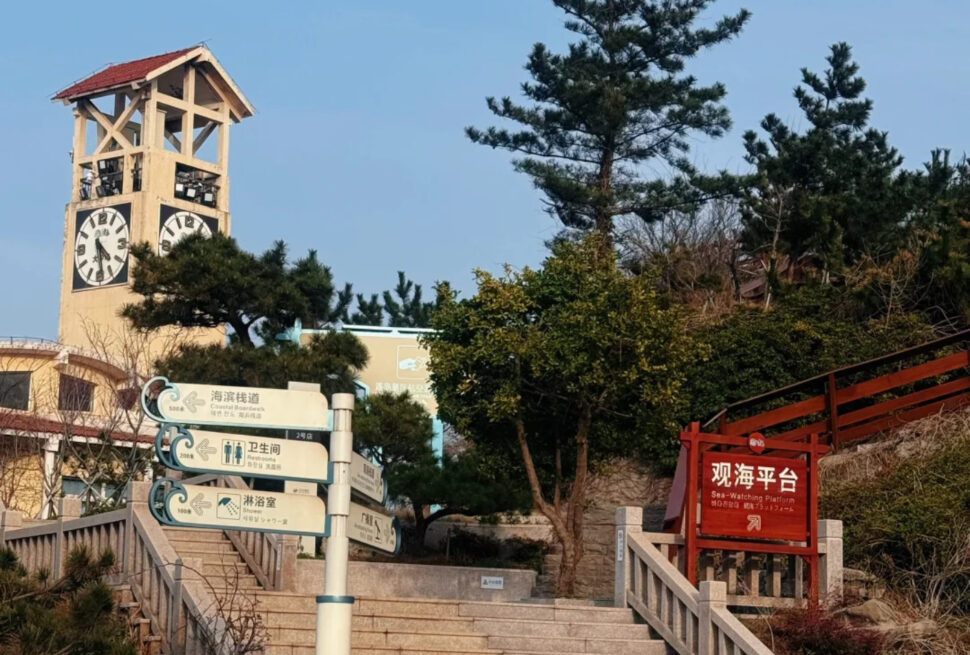 Clock tower beside a set of stone stairs with directional signs at a coastal park, blue sky above. On the right is a red-framed Sea-Watching Platform sign.
