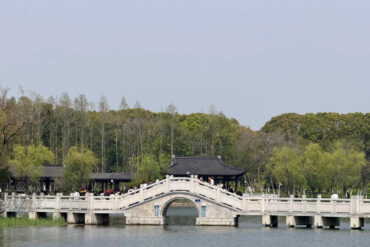 Stone arched pedestrian bridge with white railings spans a calm lake, leading to a traditional dark-roofed pavilion as visitors stroll along.