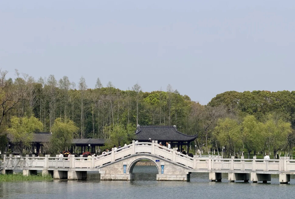 Stone arched pedestrian bridge with white railings spans a calm lake, leading to a traditional dark-roofed pavilion as visitors stroll along.