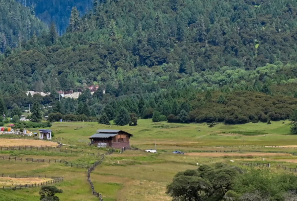 Rural valley with green fields, wooden barn, and winding fences leading toward a forested hillside and distant mountains.