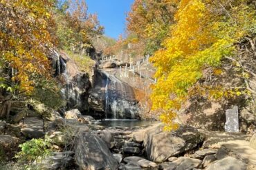 Autumn scene with golden trees framing a rocky creek and small waterfall under a clear blue sky.