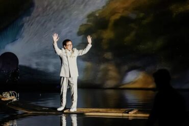 Man in a light gray suit stands on a wooden raft at night, arms raised as a colorful projection fills the backdrop behind him.