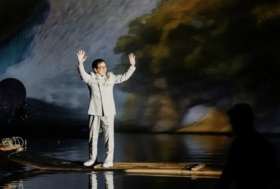 A person in traditional Chinese attire standing on a bamboo raft with arms raised on a river at nigh.