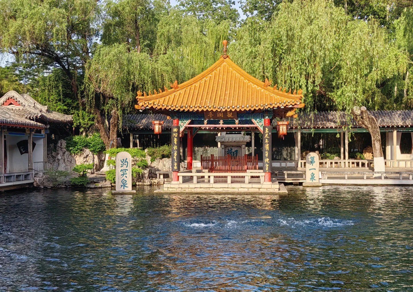 Traditional Chinese pavilion by a river with trees and lanterns.