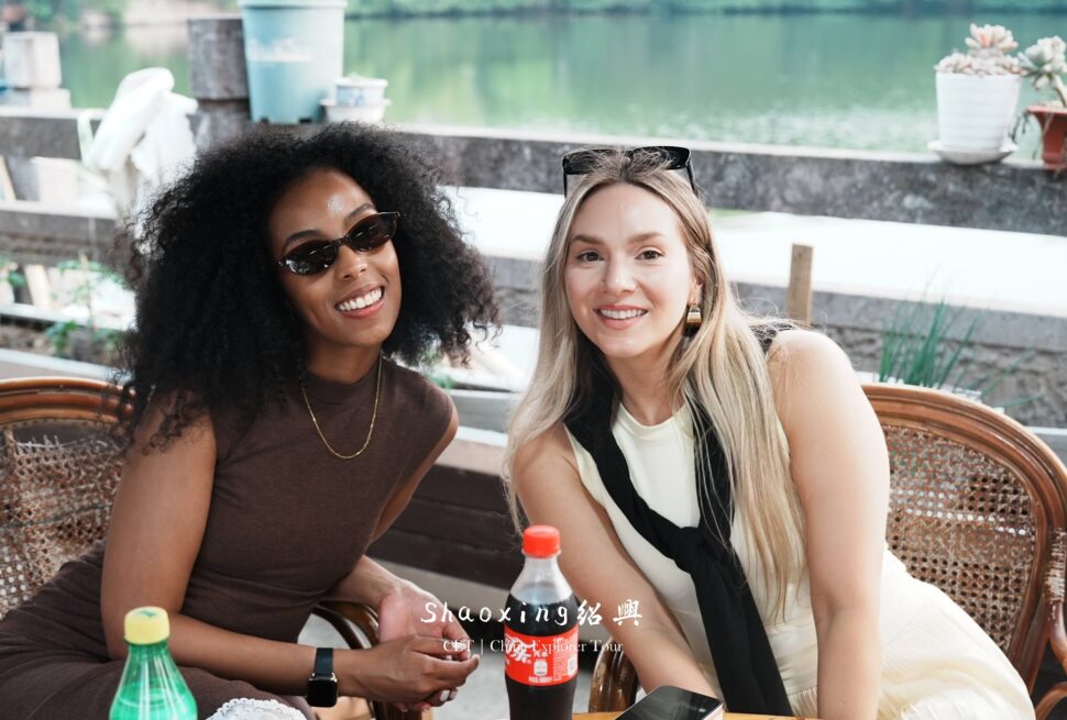 Two women smiling at a waterfront outdoor cafe with bottled drinks on the table.