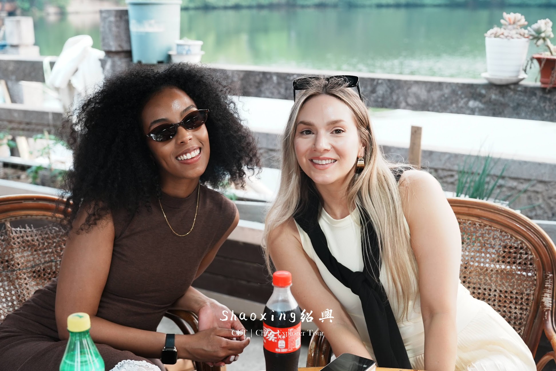 Two women smiling at a waterfront outdoor cafe with bottled drinks on the table.