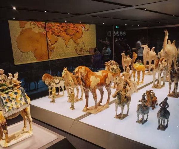Glass display case showing a caravan of terracotta figures—camels and riders—with a world map mural in the background.