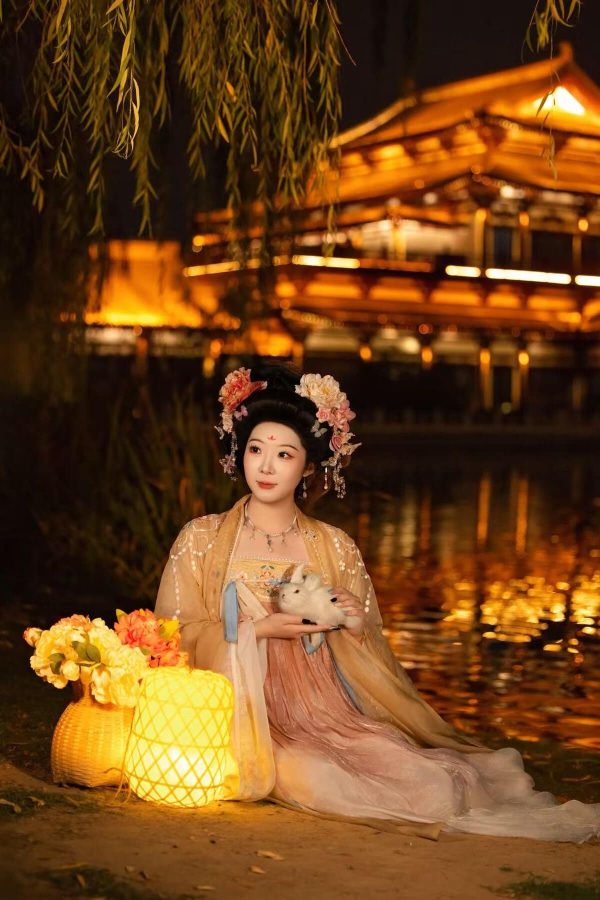 Hanfu photoshoot, xingqing palace, xi'an, solo, tang dynasty, female, chinatravel, Woman in an ornate traditional Chinese hanfu with floral hairpieces sits by a glowing lantern-lit river, holding a small white rabbit at night with a golden bridge in the background.
