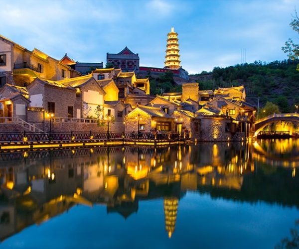 Evening view of a Chinese riverside town with lit traditional houses along a calm canal, a stone arched bridge, and a tall pagoda reflected in the water.