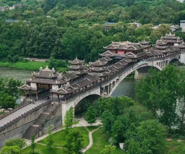 Ornate multi-pavilion Chinese bridge spanning a river, surrounded by green trees and a hillside town in the background