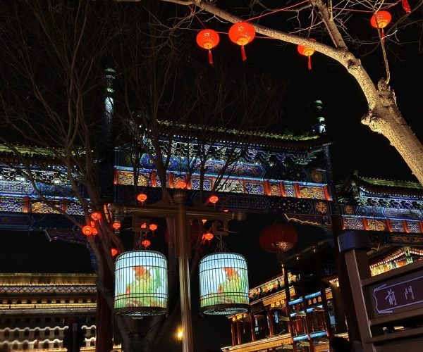 beijing, chinatravel, Night view of a traditional Chinese temple gate with red lanterns, colorful illuminated eaves, and bare trees in the foreground.