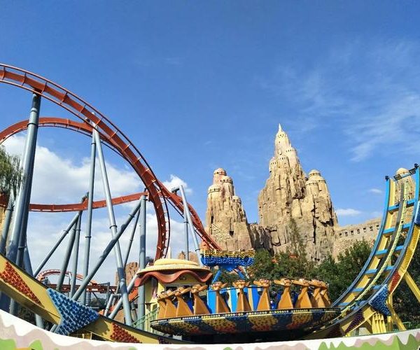 Beijing Happy Valley, Beijing, chinatravel, Colorful roller coaster with orange tracks looping around blue supports beside a rocky, castle-like structure on a sunny day at a theme park.