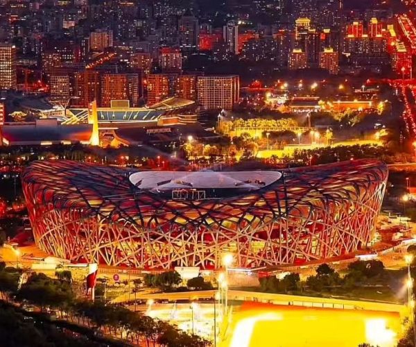 Aerial night view of the Beijing National Stadium (Bird's Nest) illuminated in orange, surrounded by city lights at dusk.