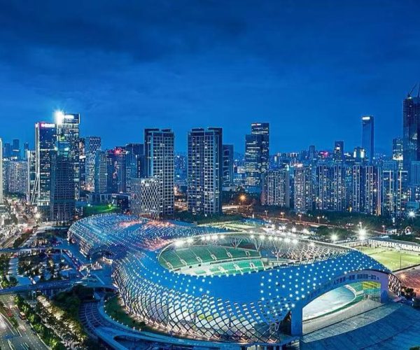 Aerial night view of a futuristic stadium with a blue lattice roof beside a dense illuminated city skyline.