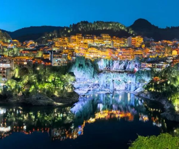 Wulong Three Natural Bridges and Furong Town,Chongqing,Chinatour Night view of a hillside town lit with warm lights, reflected in a calm river below.