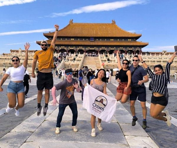 Group of friends jumping in the air in front of a large yellow-roofed Chinese palace, sunlit courtyard visible behind them, banner reads 'China Explorer'.