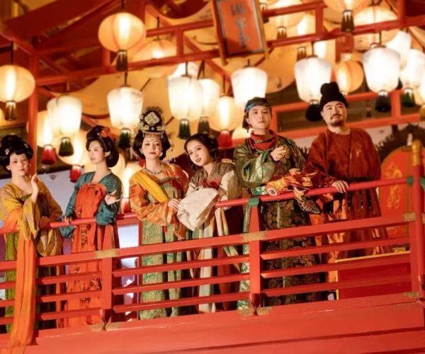 Group of people in colorful traditional Chinese costumes standing on a red balcony under hanging lanterns at a festival.