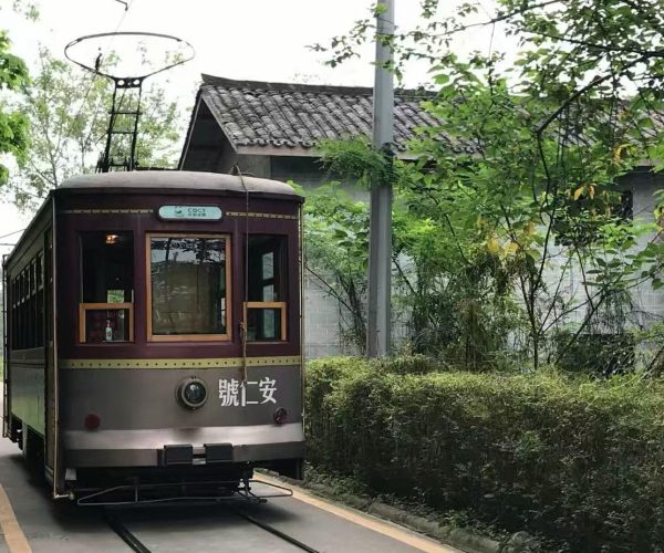 Vintage maroon tram car on a narrow rail line, flanked by trees and a hedge with a brick building in the background.