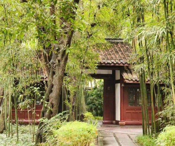 Traditional garden with bamboo, trees, and a red wooden pavilion with a tiled roof along a stone path in a tranquil courtyard.