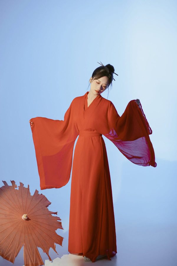 Hanfu Photoshoot Experience in China, chinatravel, female, Woman in a long orange dress with wide, flowing sleeves stands against a pale blue backdrop, arms extended outward.