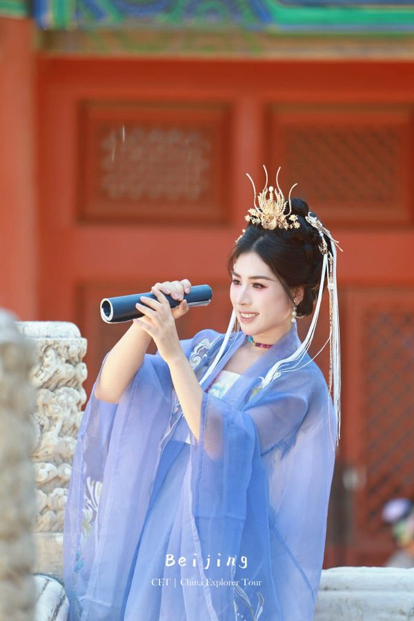Hanfu photoshoot, Beijing, Imperial Ancestral Temple (Tai Miao), female, chinatravel, Woman in a pale blue traditional Chinese hanfu with an ornate gold headdress speaks into a rolled-up microphone in a temple courtyard with red walls behind her.