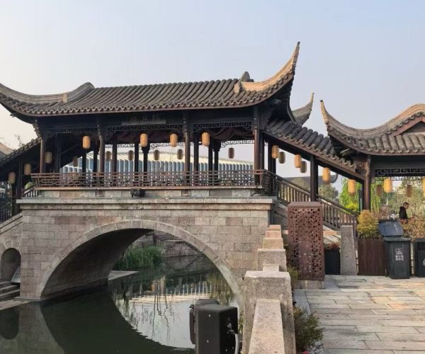 Cao'e River Pedestrian Street ,Shaoxing,Chinatour Traditional Chinese covered bridge with curved tiled roofs and hanging lanterns over a stone arch spanning a calm canal/river