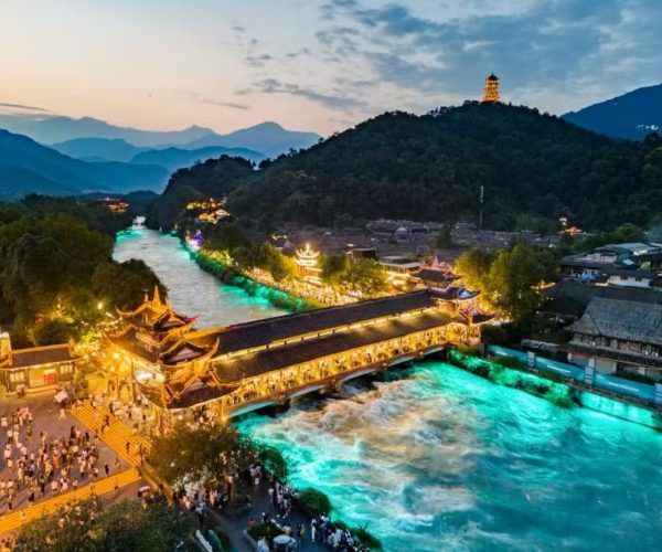 Night view of a lit temple complex over a turquoise river, with a covered bridge and a hilltop pagoda, and people on the steps.