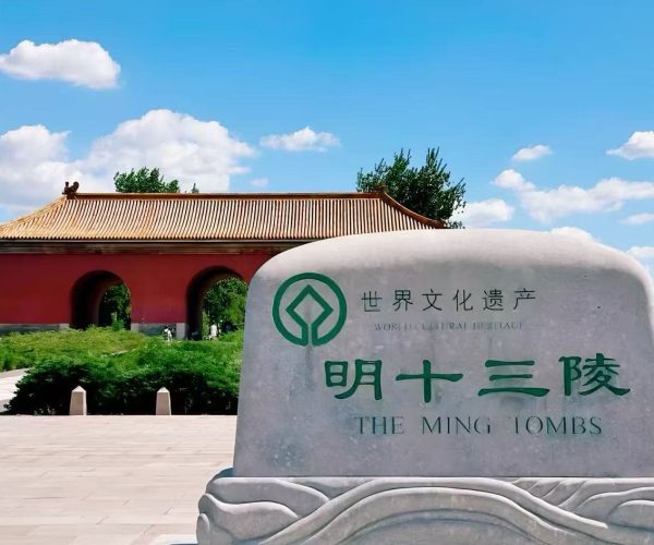 Stone monument with green emblem and Chinese text reading 'World Cultural Heritage' and 'The Ming Tombs' in front of a red gate with tiled roof under a blue sky.