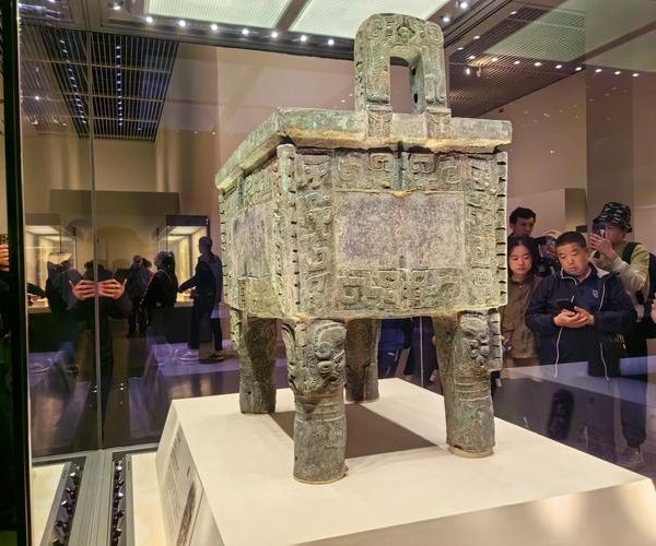 National Museum of China, beijing, chinatravel, Ancient bronze ding with four legs on display in a museum, ornate geometric patterns and a lid, visitors visible in the background behind glass shields.