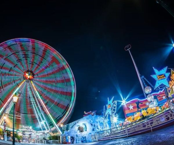 Nighttime carnival scene with a brightly lit spinning Ferris wheel and colorful rides along a illuminated promenade at a fairground edge nearby edges of the park.