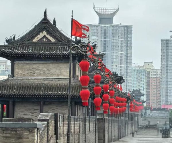 Traditional Chinese building with a curved roof and rows of red lanterns along a stone walkway, modern high-rises in the background.