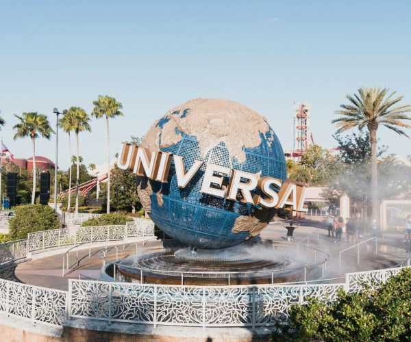 Globe sculpture with blue oceans and gold landmasses, spelling 'UNIVERSAL' in large letters, at Universal Studios theme park entrance with palm trees and mist fountain in foreground.