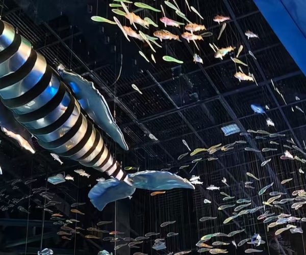 Guangzhou Science Center,Guangzhou,Chinatour Indoor aquarium scene with a large striped, spiraling hanging sculpture resembling a squid, surrounded by many small colorful fish against a dark ceiling.