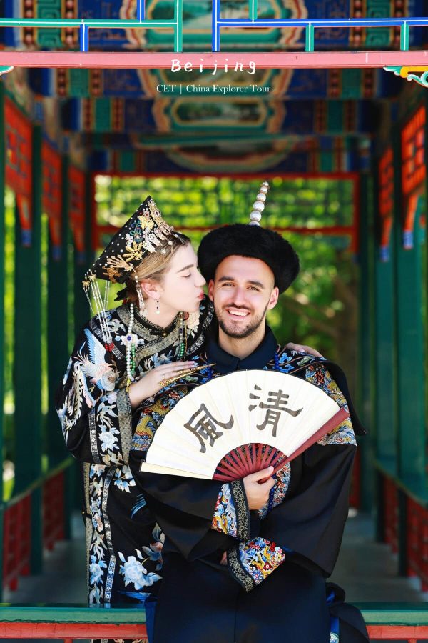 hanfu photoshoot, zhongshan park, beijing, couple, chinatrip， Couple in ornate traditional Chinese wedding outfits pose under a Beijing sign; the woman kisses the man on the cheek while holding a decorative fan.