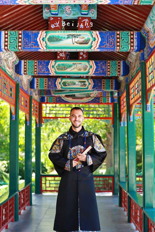 hanfu photoshoot, the forbidden city, beijing, qing dynasty, male, solo, chinatravel, Smiling man in a dark traditional Chinese robe stands in a vibrantly painted temple corridor.